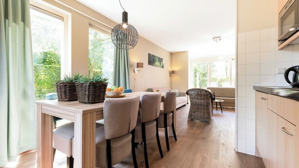 A modern kitchen with a dining area, wooden flooring, and large windows offering a view of greenery. - Comfortable bungalow for 4 persons (Diessen-Baarschot)
