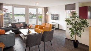 A modern living room with a sofa, armchairs, a coffee table, and a TV. There is a large window with a view of a body of water and buildings.