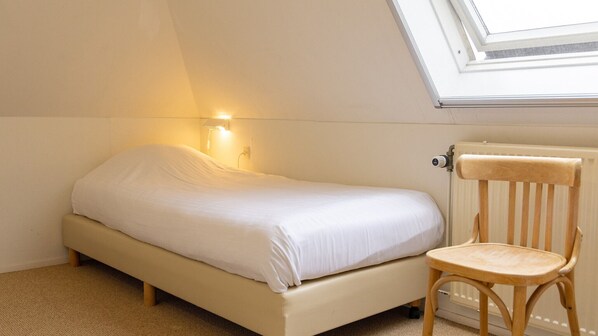 A small, single-bed room with a sloped ceiling and a skylight. There is a wooden chair and a radiator on the wall.