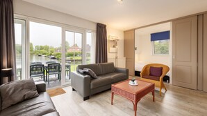 Een ruime woonkamer met een grijze bank, een rieten salontafel en een rieten fauteuil. Grote ramen bieden uitzicht op een gracht en boten.