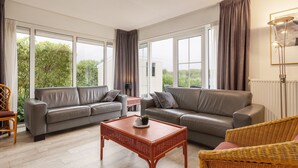 Een ruime woonkamer met twee leren banken, een houten salontafel, rieten stoelen en grote ramen met uitzicht op groen.