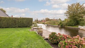 Un canal con una embarcación amarrada a la derecha, una zona verde con una mesa y sillas de madera, y un seto a la izquierda.