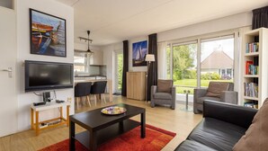 Una sala moderna con una televisión de pantalla plana, un comedor con sillas, un sofá, una estantería y vistas a una casa a través de amplios ventanales.