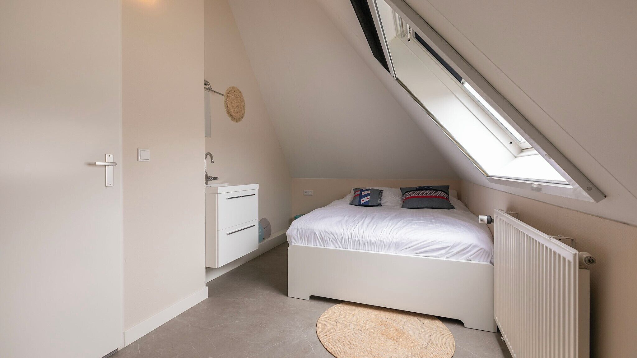 A small, white, sloped-roof bedroom with a bed, a radiator, a window, and a door.