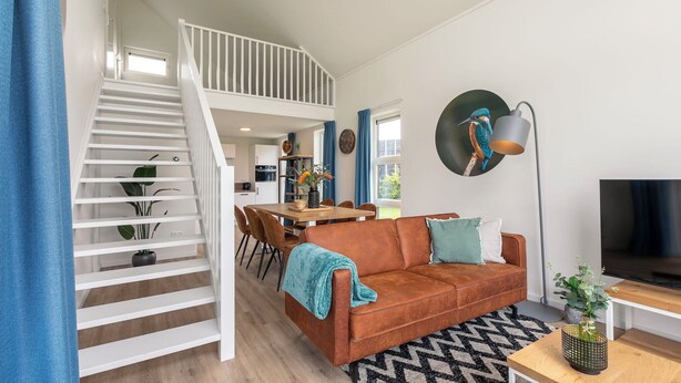A modern apartment with a staircase leading to a loft, a dining area, a living room with a sofa and TV, and a kitchen in the background. - Detached villa for 6 persons (Tynaarlo)