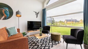 A modern living room with a large TV, comfortable seating, a patterned rug, and a view of the outdoors. - Detached villa for 6 persons (Tynaarlo)