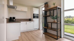 A modern kitchen with white cabinetry, a built-in oven, and a coffee maker. There is a wooden shelf unit with plants and a round wooden object on top. - Detached villa for 6 persons (Tynaarlo)