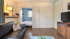 A compact living space with a leather sofa, a TV on a wooden stand, and a bed in the background.