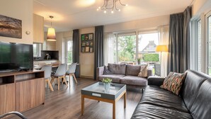 A modern living room with a sofa, coffee table, and TV. There is a kitchen area with a dining table and chairs. Large windows provide natural light and views of the outdoors.