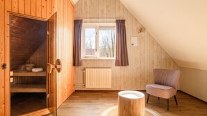 A cozy attic room with wooden walls, a window with curtains, a radiator, a chair, and a small table. - Luxury bungalow for 4 people (Diessen-Baarschot)