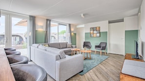 A modern living room with a grey sectional sofa, two armchairs, a coffee table, and a flat-screen TV. There are large windows with curtains, a green accent wall, and a view of the outside.
