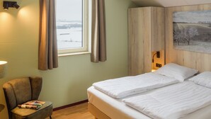 Habitación de hotel con cama, una silla, ventana con cortinas, lámpara y un cuadro en la pared.