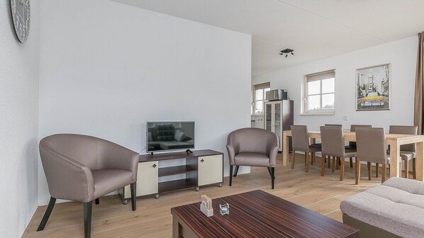 A modern living room with a flat-screen TV, two leather armchairs, a wooden coffee table, and a dining area with a wooden table and chairs.