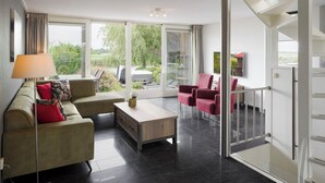 Un salon moderne avec un canapé, une table basse et des fauteuils, de grandes fenêtres offrant une vue sur la verdure et un jacuzzi à l’extérieur.