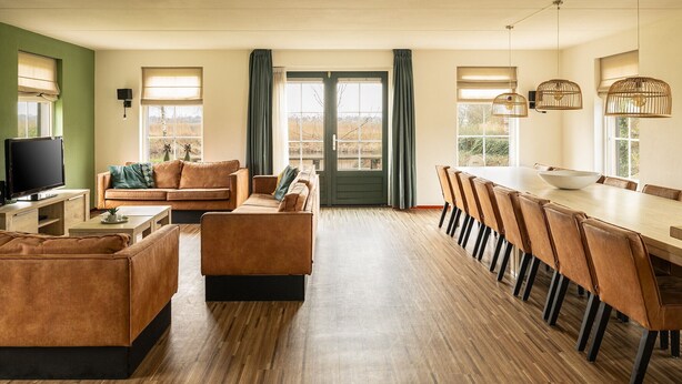 A spacious living area with a sofa, coffee table, and TV, leading to a dining area with a long table and chairs. - Bungalow Wellness 16 (Vlagtwedde)