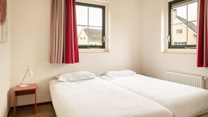 A bedroom with two beds, a nightstand, a lamp, and two windows with red curtains. - Bungalow Wellness 16 (Vlagtwedde)