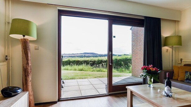 Een kamer met een schuifdeur naar een terras, een houten tafel met een bloevas en twee lampen.