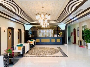 Un hall d’hôtel comprenant un comptoir d’accueil, un lustre, une moquette à motifs et un espace détente avec des tables et des chaises.