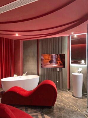 Une salle de bain moderne avec une grande baignoire sur pieds, un banc recouvert d’un tissu rouge, un grand miroir et un téléviseur à écran plat fixé au mur.