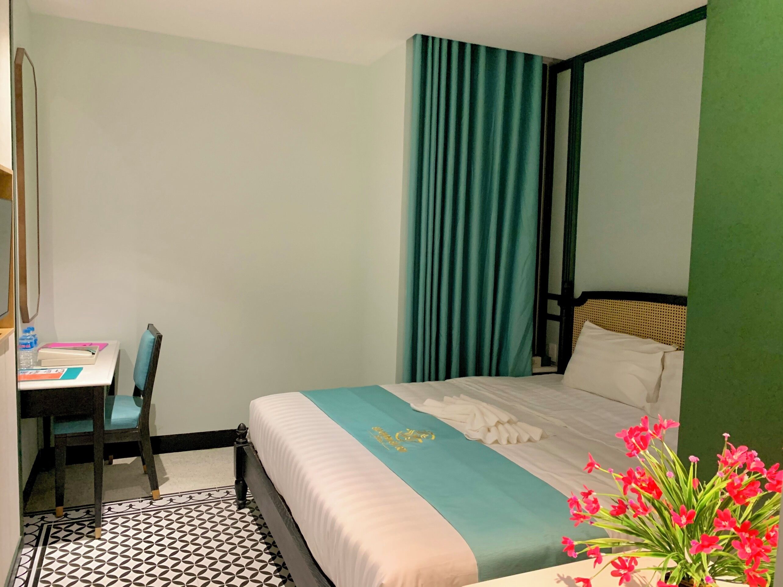 A hotel room with a bed, a desk with a chair, a mirror, and a potted plant with flowers.