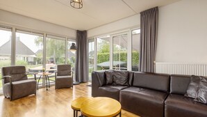 Un salon spacieux, doté d’un canapé en cuir, de deux fauteuils et d’une table basse en bois. De grandes fenêtres offrent une vue sur l’extérieur.
