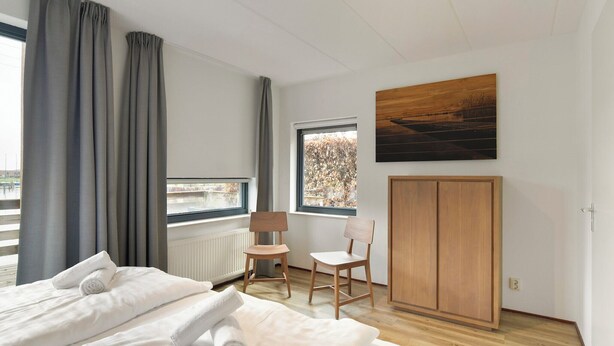A modern bedroom with a bed, two chairs, a wardrobe, and a window with a view of trees. - Comfortable bungalow for 6 persons at the waterfront (Midlaren)
