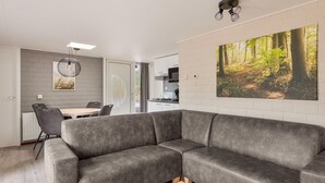 A modern living room with a grey sectional sofa, a dining area with a table and chairs, a kitchen with white cabinets, and a large forest-themed painting on the wall. - Semi-detached bungalow for 2 persons (Kootwijk)