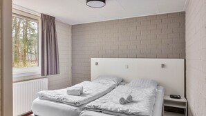 A bedroom with two beds, a window with curtains, a brick-patterned wall, and a white headboard. - Semi-detached bungalow for 2 persons (Kootwijk)