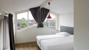 Ein Schlafzimmer mit zwei Betten, einem fensterbelaubten Fenster, einer roten Pendelleuchte und Blick auf Gebäude.