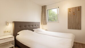 A bedroom with a double bed, a bedside table, a nightstand, a wall-mounted light, a window with curtains, and a wooden panel on the wall.