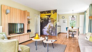 Un salon moderne doté d’un téléviseur à écran plat, de lambris en bois, d’un coin repas avec une table et des chaises, et d’une cuisine avec des meubles blancs.
