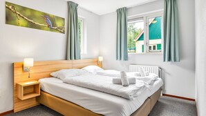 Une chambre d’hôtel avec une tête de lit en bois, deux tables de chevet avec des lampes, un grand lit avec du linge de lit blanc et une fenêtre avec des rideaux verts.
