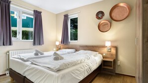 Cama doble con ropa blanca, dos mesitas de noche con lámparas, cabecera de madera y una ventana con cortinas.