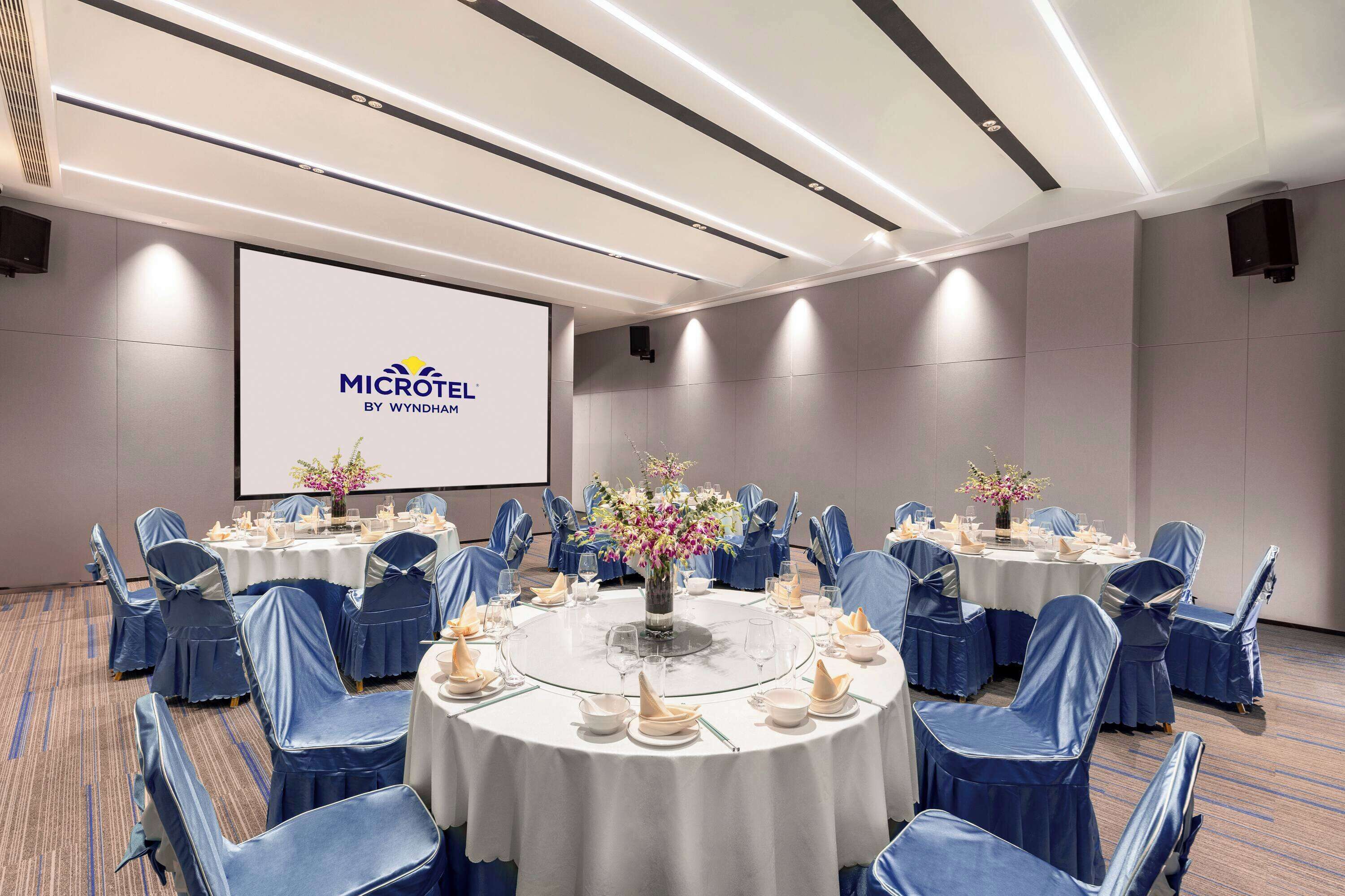 Ett konferensrum med runda bord, stolar klädda i blått tyg och en stor skärm som visar MICROTEL BY WYNDHAM-logotypen.