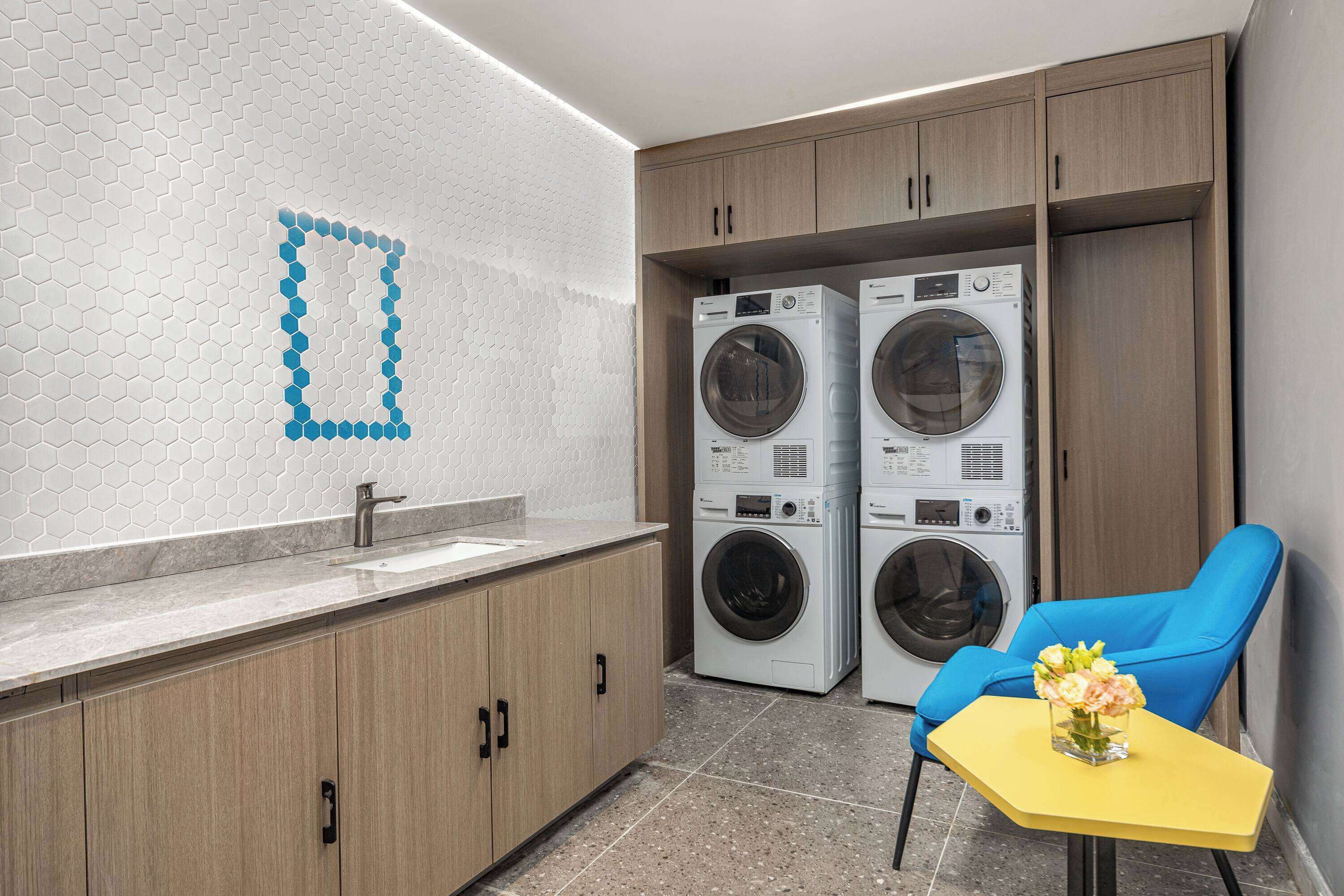 A modern laundry room with two washing machines, a sink, wooden cabinets, and a blue chair with a vase of flowers.