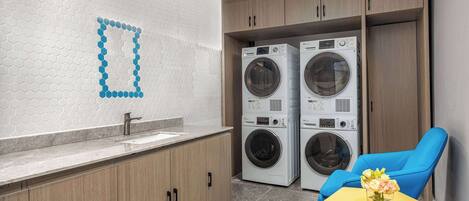A modern laundry room with two washing machines, a sink, wooden cabinets, and a blue chair with a vase of flowers.
