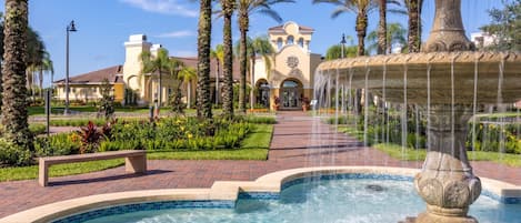 Una fuente con chorros de agua, una piscina, palmeras y un edificio con tejado de baldosas al fondo.