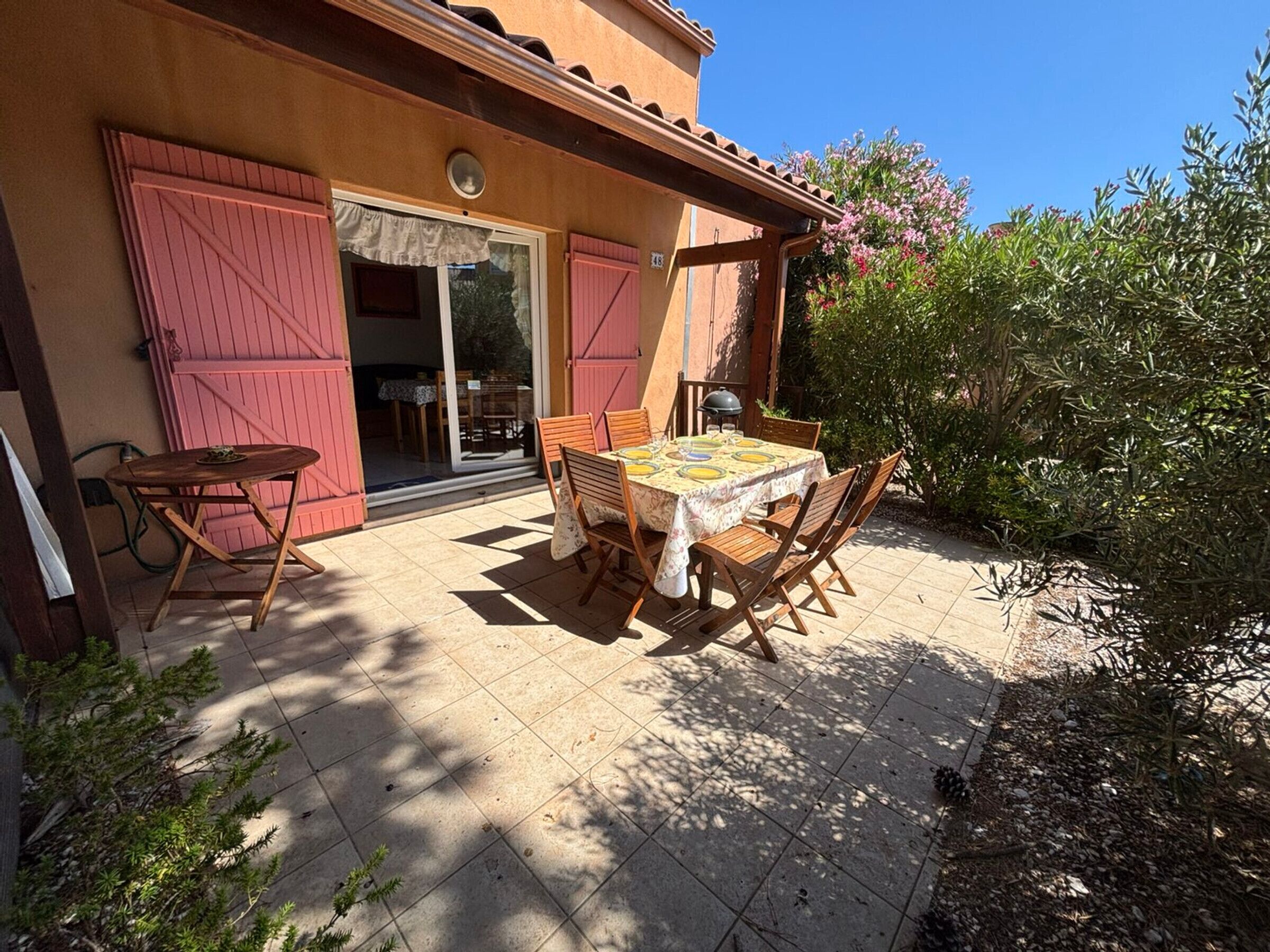 Un patio con tavolo e sedie, una porta scorrevole in vetro e un giardino con arbusti e fiori.