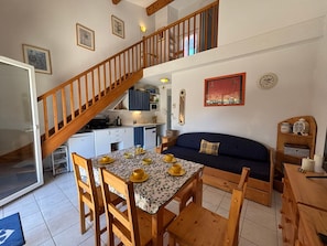 Ein Essbereich mit einem Tisch fĂŒr vier Personen, eine Holztreppe, eine KĂŒche und ein Wohnbereich mit einer Couch.