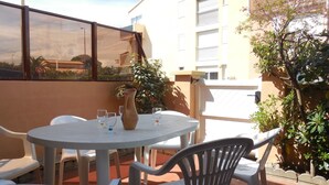 A round table with chairs and a vase, set on a terrace with a view of a building and a tree.