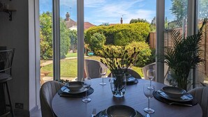 Un comedor con una mesa dispuesta para cuatro personas, amplios ventanales que ofrecen vista a un jardín cuidado y un edificio lejano.