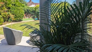 Un patio con una silla de mimbre y una planta grande en maceta, con vista a un jardín cuidado y un edificio lejano.