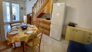 A kitchen with wooden cabinets, a white refrigerator, and a dining table set with plates and a bowl. - T2 Mezzanine Villa - Air Conditioning - Veranda - Parking - Pool - 6 People (Fleury-Saint-Pierre-la-Mer)