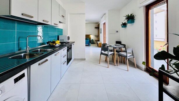 Une cuisine moderne avec des armoires blanches, un comptoir noir et un pare-feu turquoise. On y trouve un coin repas avec une table et des chaises, ainsi qu’une fenêtre ornée de plantes sur le rebord.