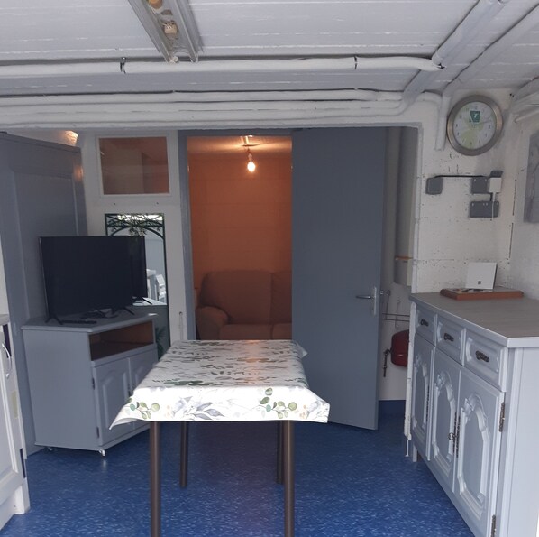 Une petite pièce avec un téléviseur posé sur un meuble, une table recouverte d’une nappe à motifs floraux et une porte donnant sur une autre pièce.