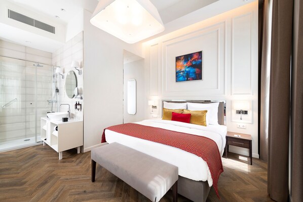 Una habitación de hotel moderna con una cama grande, una mesita de noche, un banco, un baño con ducha de vidrio y un cuadro en la pared.