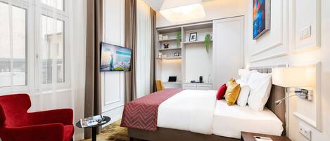 A hotel room with a bed, a red armchair, a small table, a TV, a desk, and a bookshelf.
