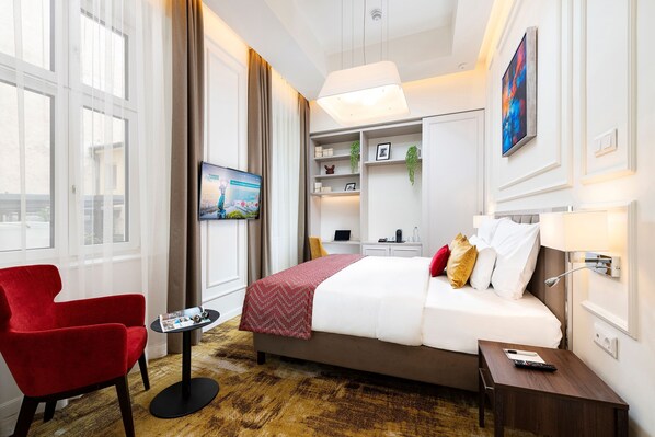 Una camera d'albergo con un letto, una poltrona rossa, un tavolino, una TV, una scrivania e una libreria.