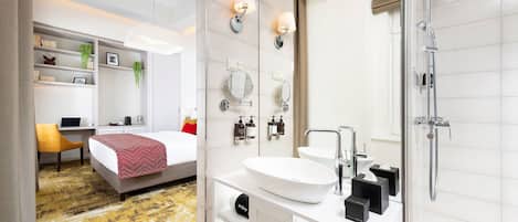 A modern hotel room with a large bed, a desk with a laptop, a white sink, and a glass-enclosed shower.