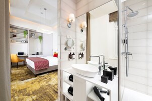 A modern hotel room with a large bed, a desk with a laptop, a white sink, and a glass-enclosed shower.